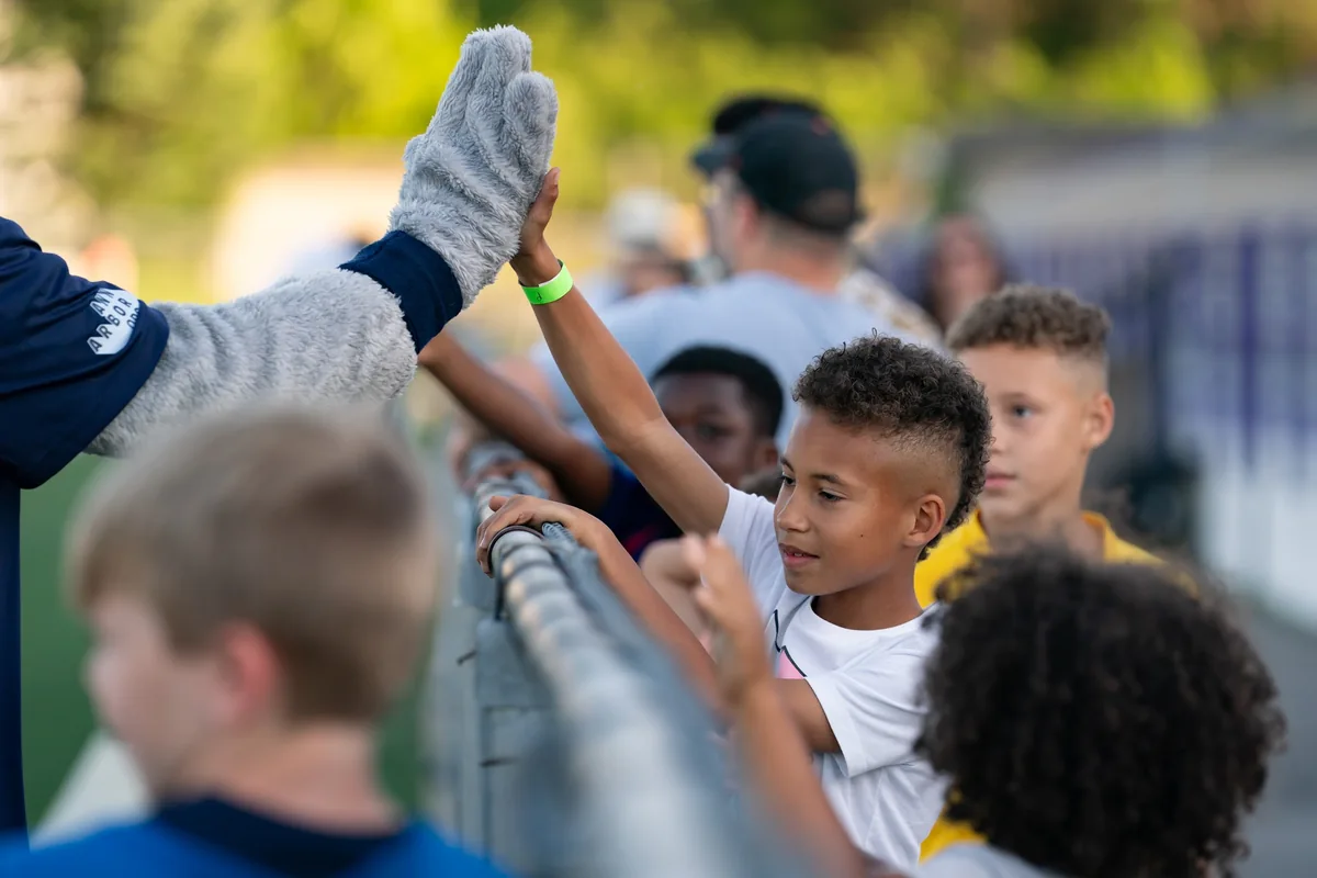 A young supporter high-fiving the club mascot at the pitch-side fence.