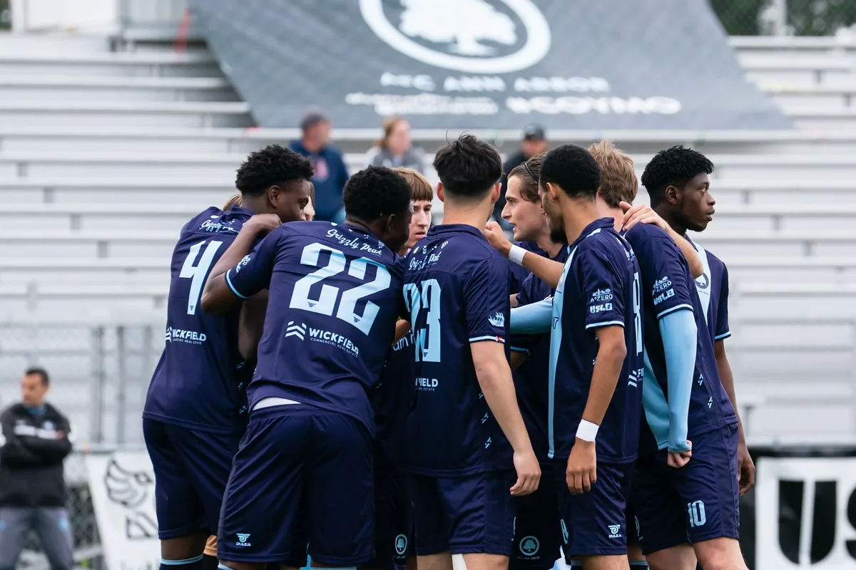 AFC Ann Arbor men’s players in navy and sky blue kits, with their arms around one another in a team huddle.