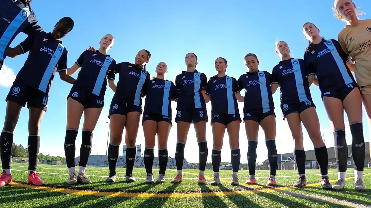 AFC Ann Arbor women’s team in navy and sky blue kits, standing together in a huddle beneath a blue sky.